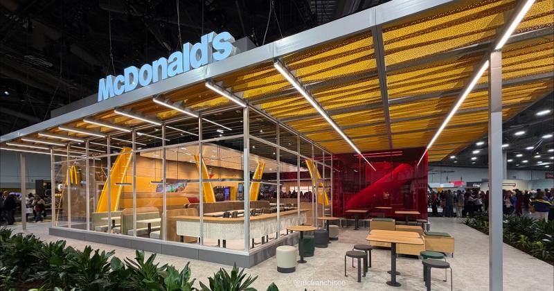 McDonald's new PlayPlace. Modern McDonald’s restaurant with glass walls, yellow accents, and outdoor seating. People are visible inside and outside the building. The setting appears to be indoors, possibly at an event or expo.