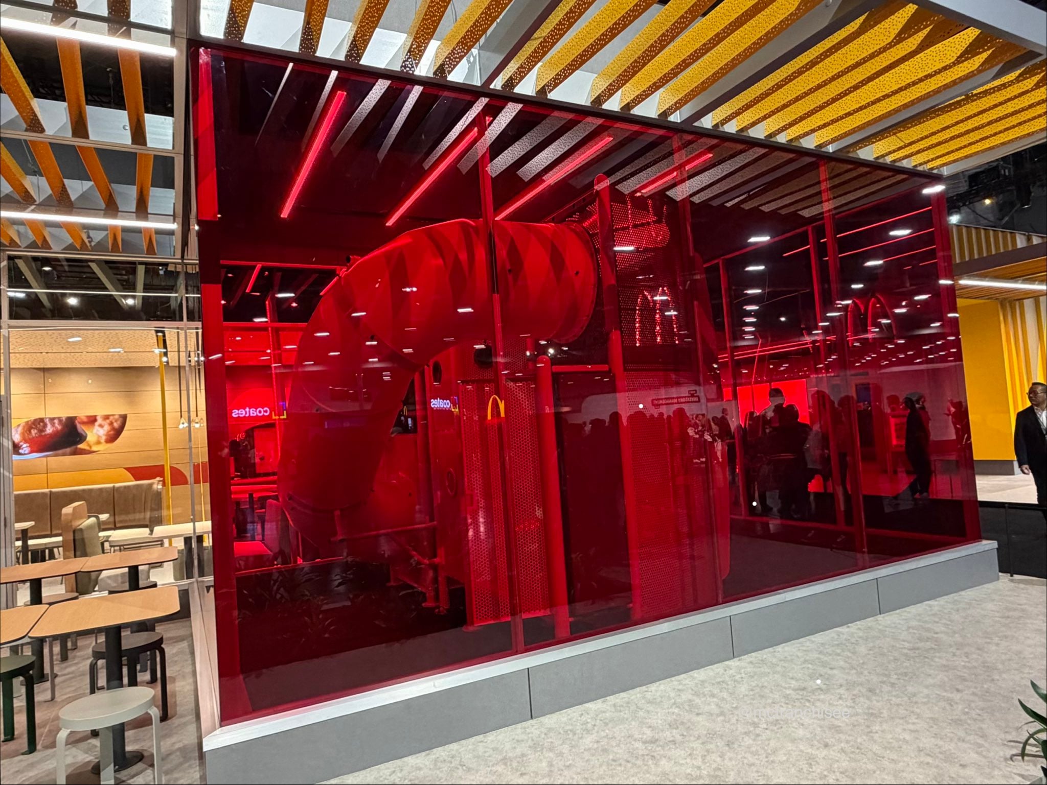 A large red glass wall reveals a kids’ play area with a spiral slide inside a modern fast food restaurant, surrounded by tables and seating.