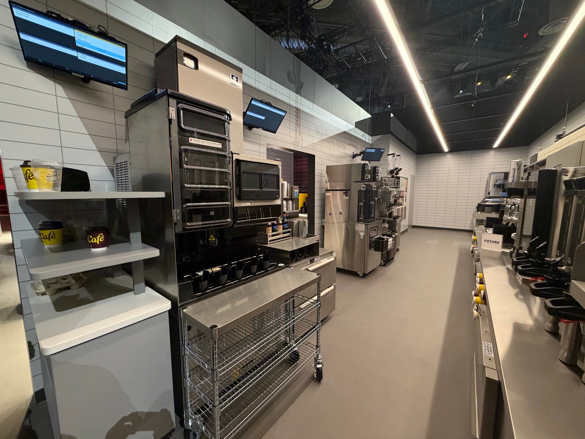 A modern commercial kitchen with stainless steel appliances, shelves, ovens, and two wall-mounted monitors displaying digital screens. The room is brightly lit with overhead lights and has a clean, organized appearance.