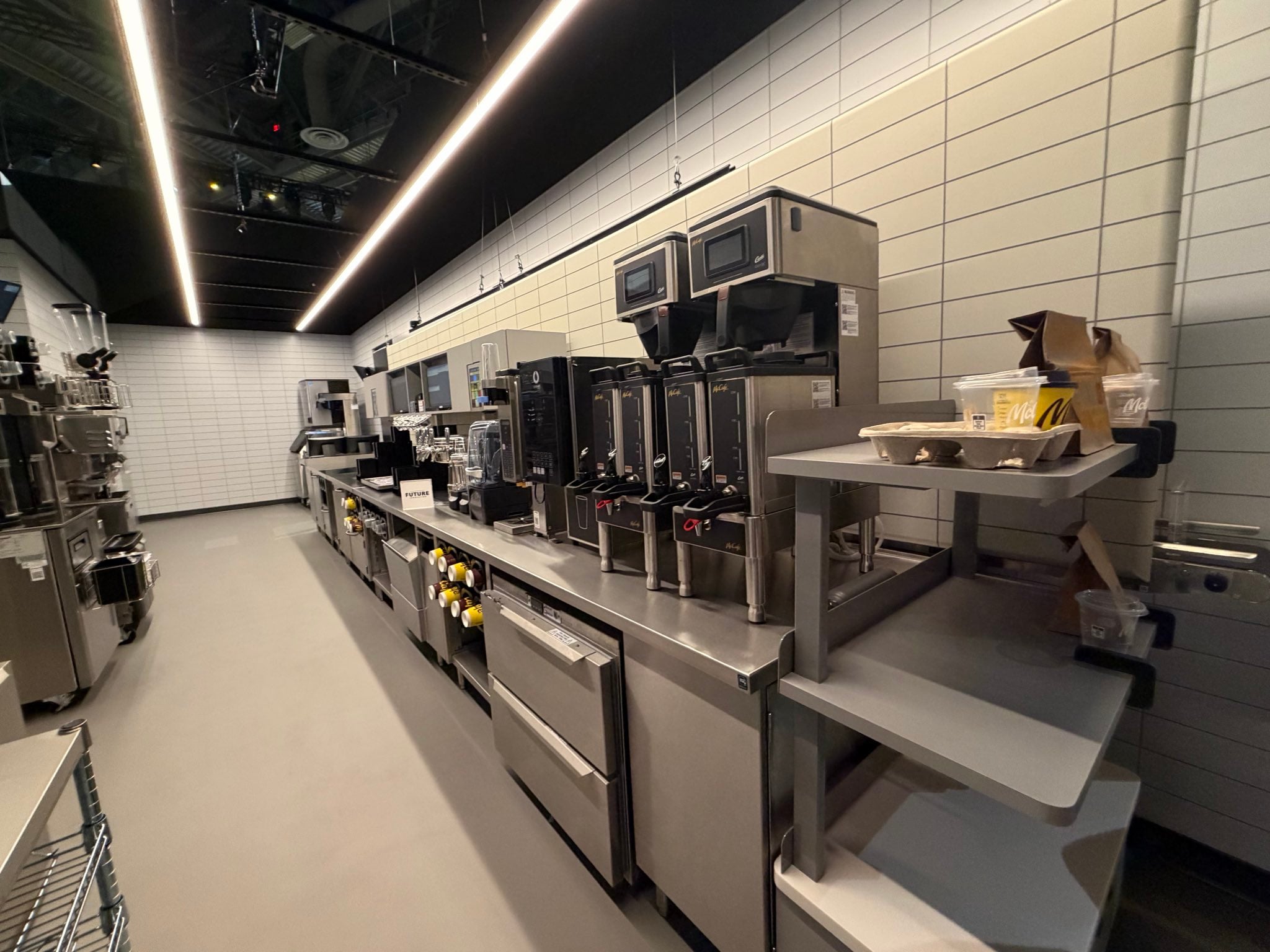 A modern commercial kitchen with stainless steel equipment, including coffee machines, ovens, and shelves. The walls are tiled white, and the space appears clean and organized.