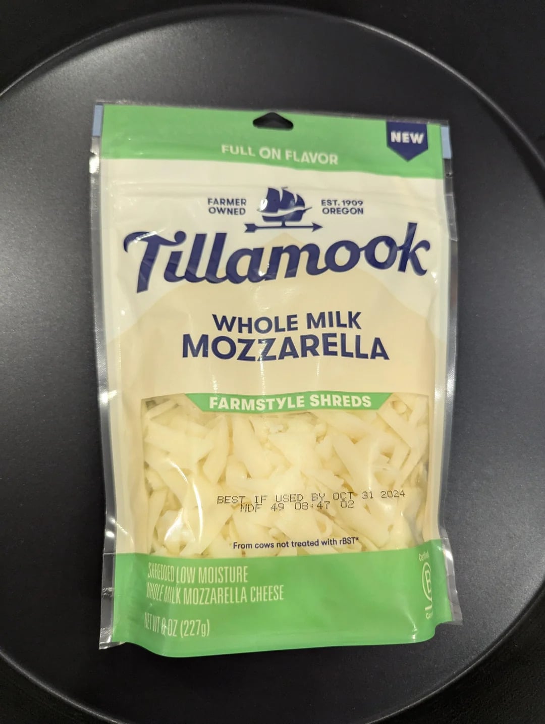 A bag of Tillamook Whole Milk Mozzarella Farmstyle Shreds, known as one of the best shredded cheese options, sits on a dark surface. The package is mostly white and green, and displays "Full On Flavor" and "Farmer Owned" at the top.