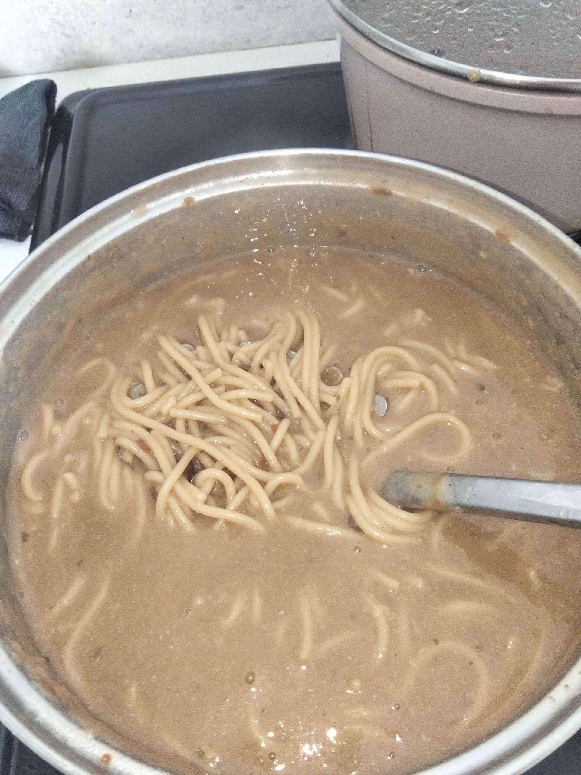 A pot of cooked spaghetti noodles in a thick, brown sauce is being stirred with a metal spoon on a stovetop. A second pot with a lid is visible in the background.