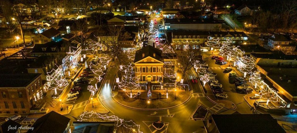 Aerial view of a town square lit with holiday lights at night, featuring a central building surrounded by trees wrapped in string lights and nearby streets glowing with festive decorations—capturing the magical charm found in some of the best Christmas towns in America.