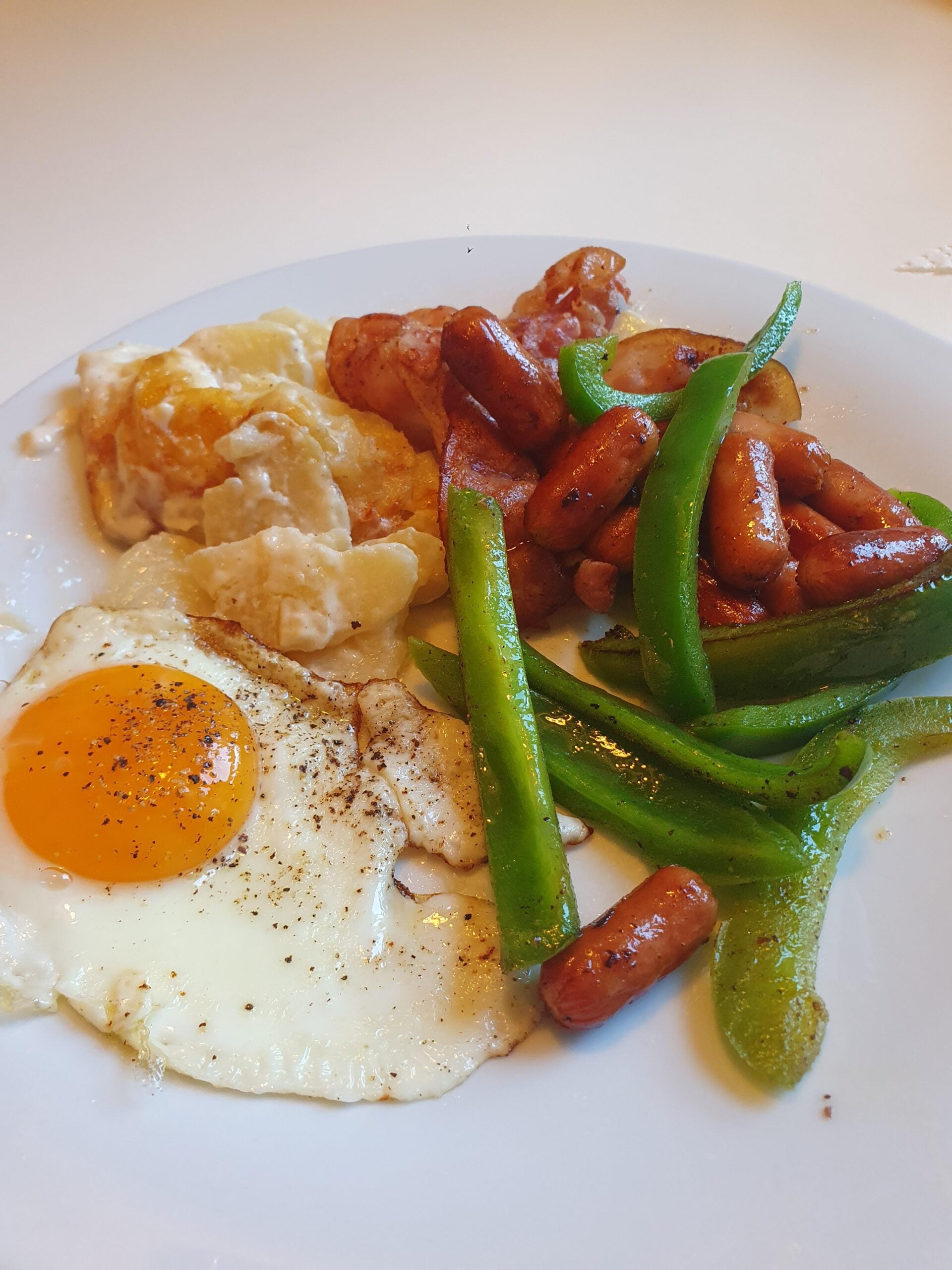 A plate with a sunny-side-up egg, sautéed green bell peppers, mini sausages, and creamy potato gratin, arranged on a white surface.
