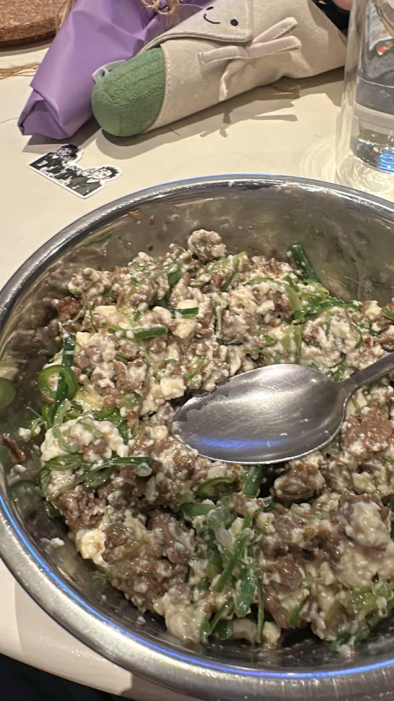 A metal bowl filled with a mixed salad of meat, green herbs, and cheese, with a large metal spoon inside. In the background are a glass of water, a plush toy, and a napkin.