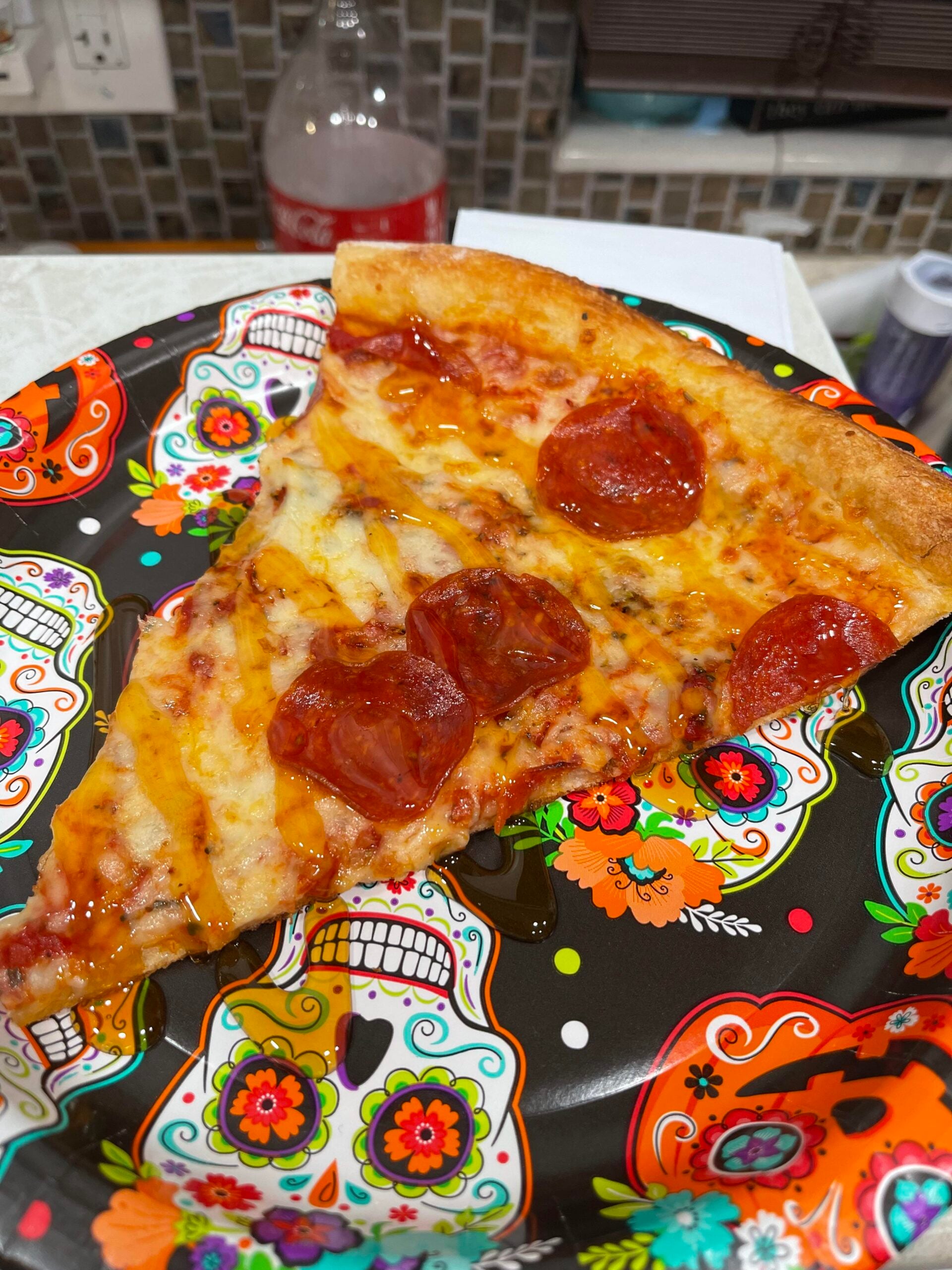 A slice of pepperoni pizza on a colorful decorative plate with sugar skull designs, with a soda bottle and kitchen items blurred in the background.