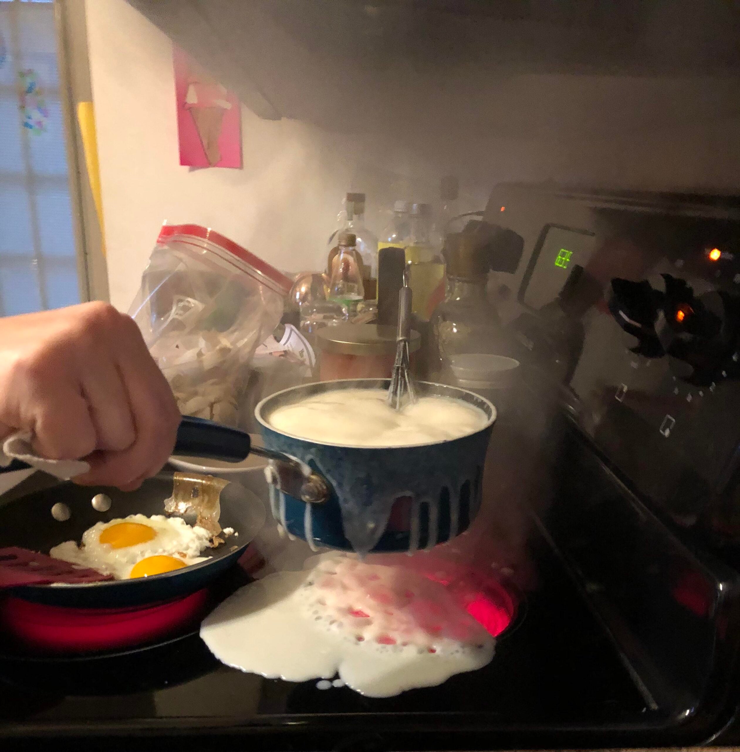 A pot of milk boils over onto a stovetop while someone lifts it, capturing one of those hilarious cooking disasters. Two eggs fry in a pan beside the overflowing pot, with various kitchen items visible in the background.