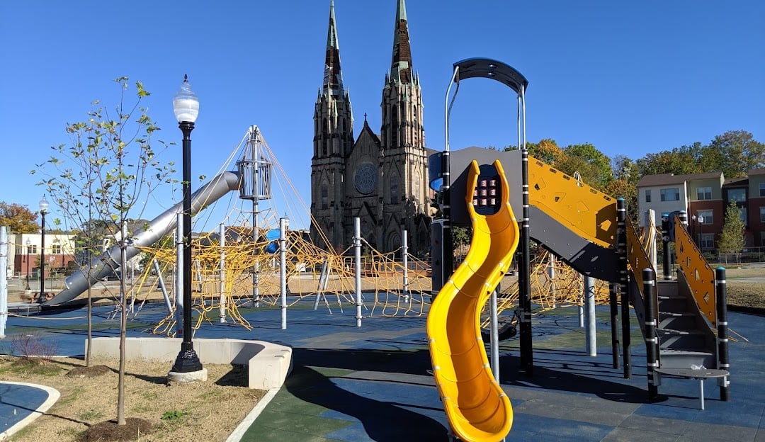A modern playground with slides and climbing structures sits in front of a large, historic church with two tall spires under a clear blue sky. Young trees and a lamppost are also visible.