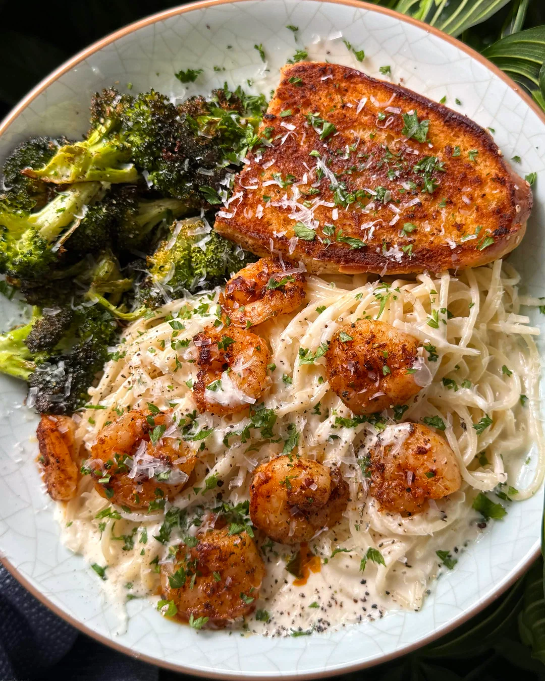 A plate with creamy shrimp pasta, roasted broccoli, and a slice of toasted garlic bread, garnished with grated cheese and chopped herbs.