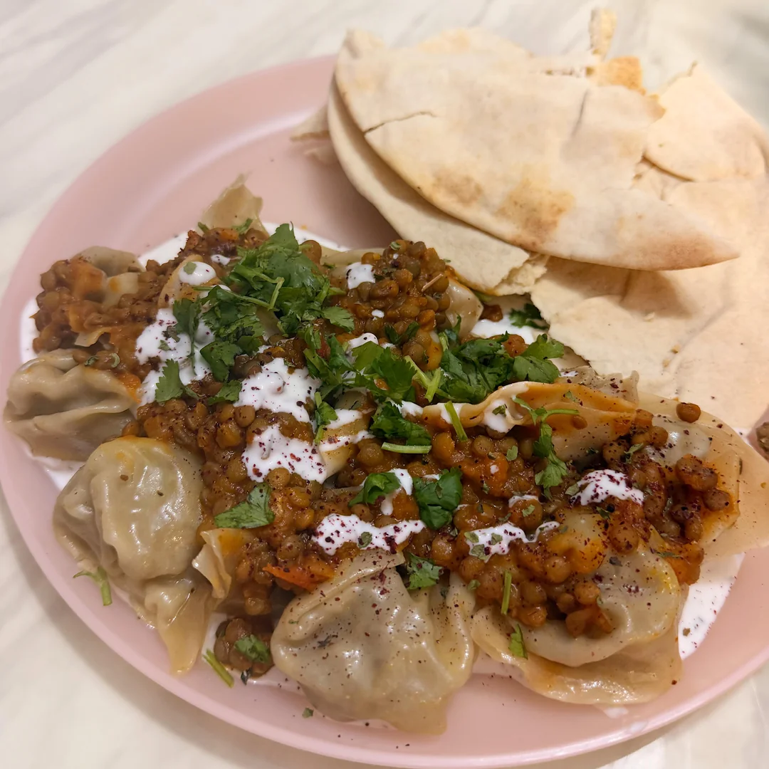A plate with dumplings topped with lentils, sauce, and chopped herbs, served alongside pieces of pita bread on a light pink plate.