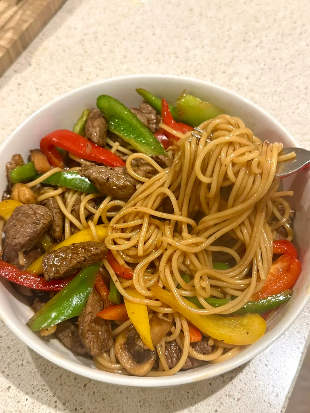A white bowl filled with stir-fried noodles, beef strips, and colorful bell peppers sits on a countertop. A fork lifts a portion of noodles above the bowl.