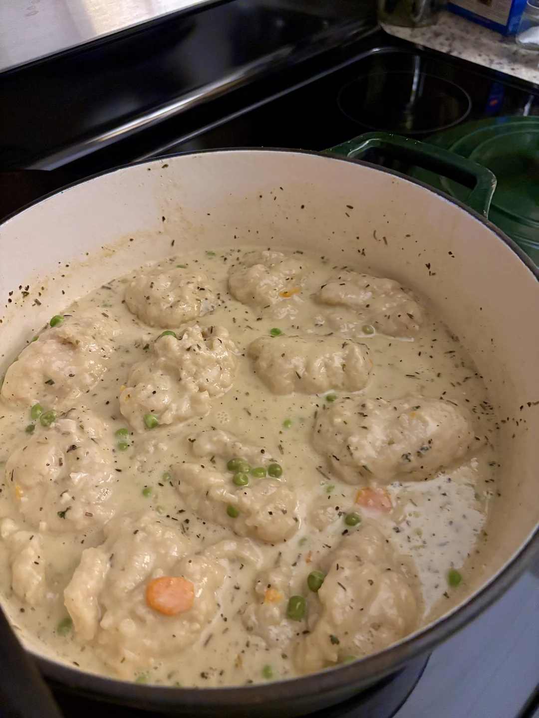 A large pot of creamy chicken and dumplings, with biscuit-like dumplings, peas, carrots, and herbs simmering in a thick, savory broth on a stovetop.