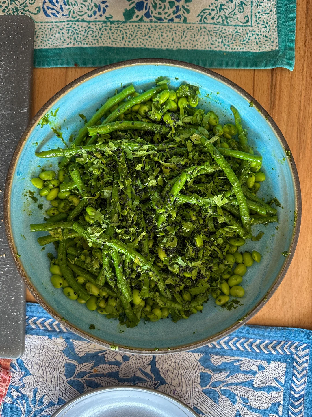 A blue bowl filled with green beans, edamame, and chopped herbs sits on a patterned tablecloth, next to a cutting board.