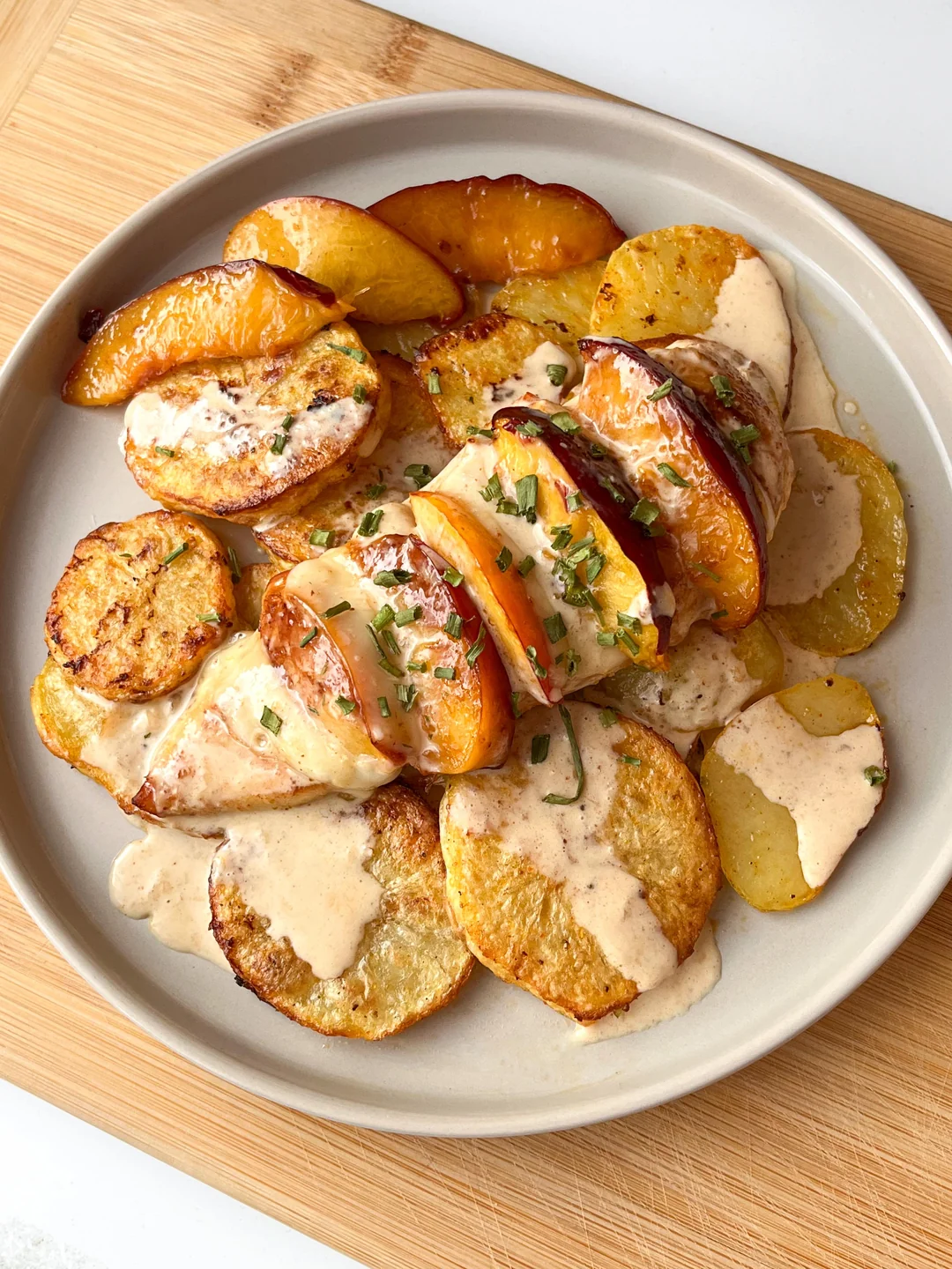 Sliced roasted potatoes and peaches arranged on a plate, topped with creamy sauce and chopped herbs, served on a light gray dish over a wooden board.