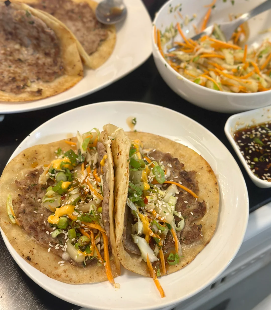 Two folded tacos filled with vegetables and sauce are on a white plate. Behind them are more flatbreads with meat, a bowl of shredded veggies, and a dish of dark dipping sauce. Everything is arranged on a table.