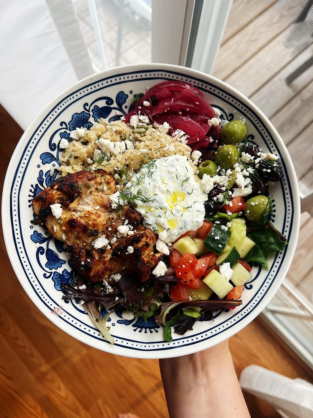 A hand holds a plate with grilled chicken, brown rice, tzatziki, pickled onions, olives, feta, cucumber, tomato, and greens, beside a window with sunlight streaming in.