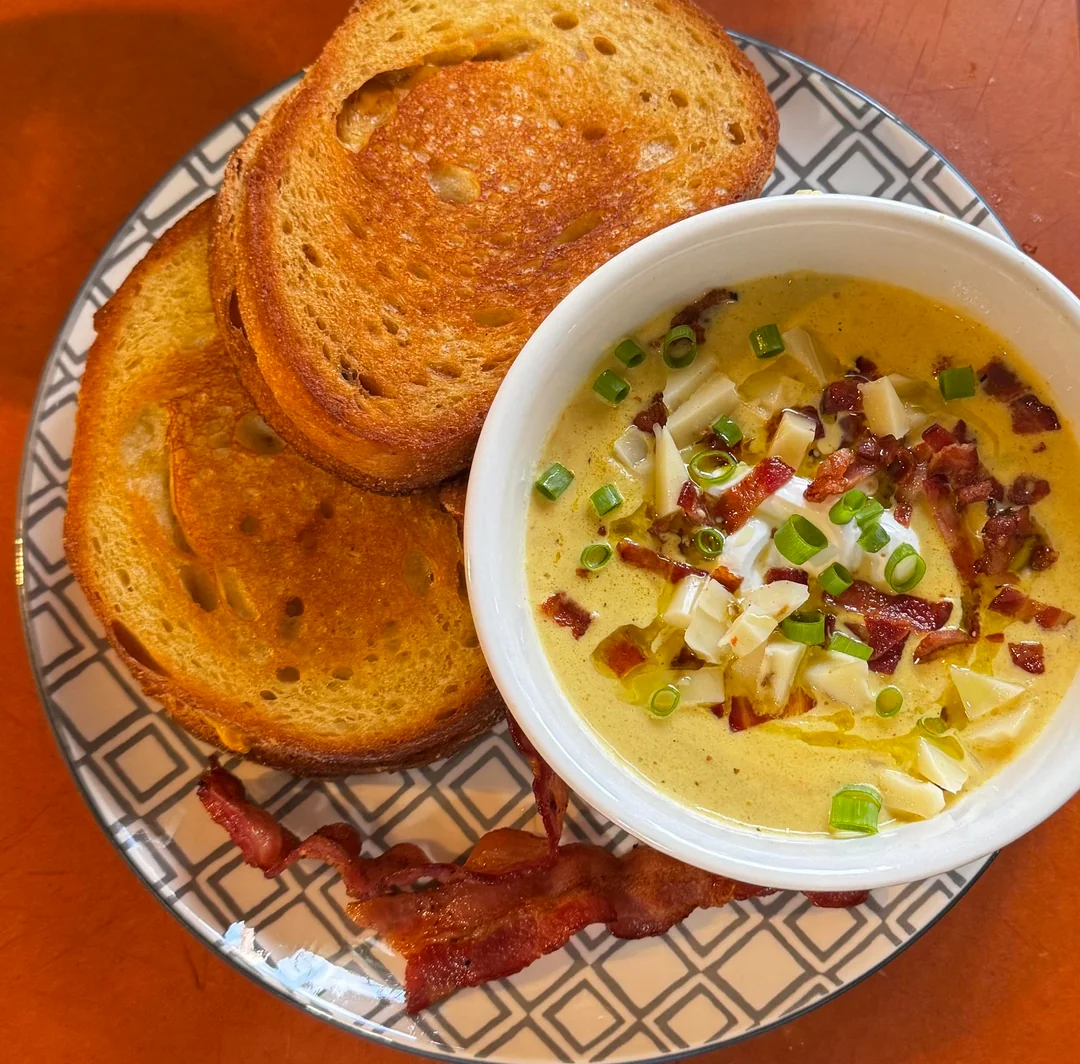 A bowl of creamy soup topped with chopped bacon, green onions, and cheese sits on a plate with two slices of toasted bread and a strip of bacon. The plate has a geometric pattern.