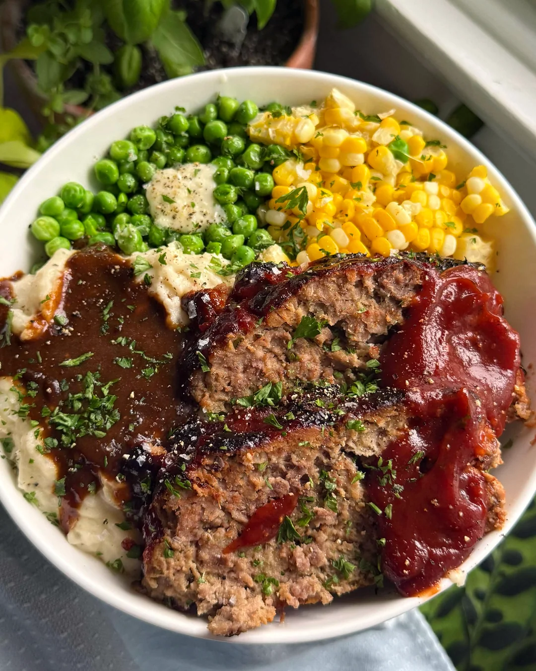 A bowl with mashed potatoes and gravy, green peas, corn, and two slices of glazed meatloaf, garnished with chopped parsley.