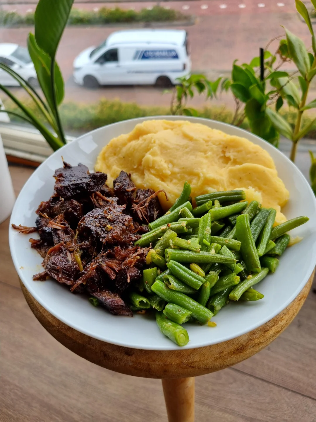 A plate with mashed potatoes, green beans, and shredded beef sits on a wooden stool by a window with plants and a white van visible outside.