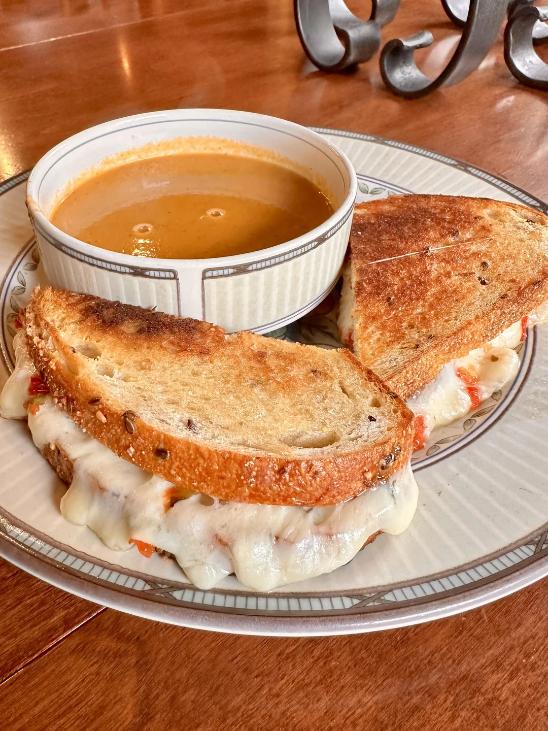 A bowl of tomato soup sits on a plate with two halves of a grilled cheese sandwich made with multigrain bread and melted cheese, resting beside it on a wooden table.