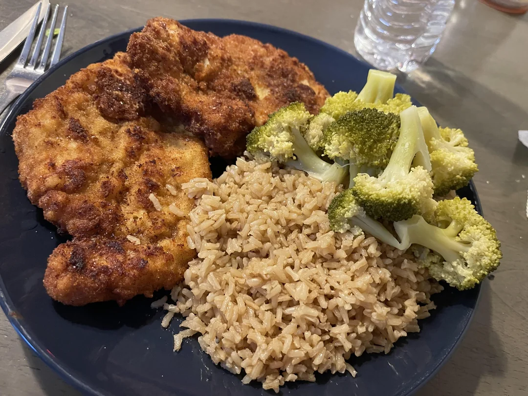 A blue plate with breaded chicken cutlets, a serving of brown rice, and steamed broccoli. A water bottle and part of a fork are visible in the background.