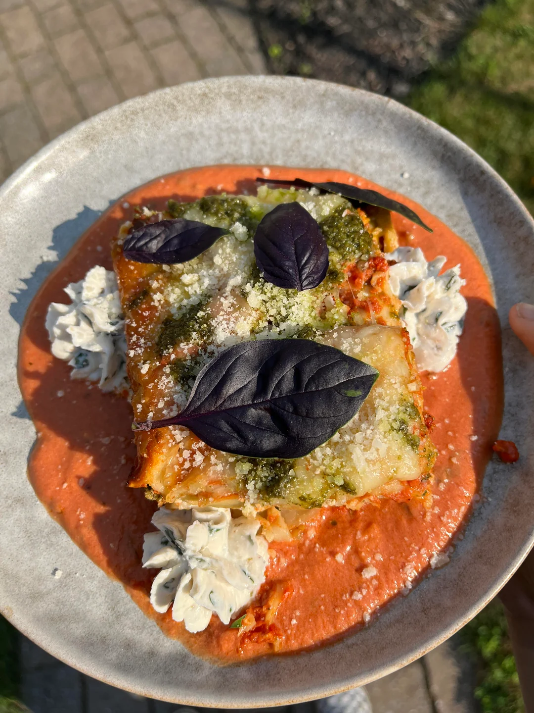 A slice of lasagna garnished with dark basil leaves and dollops of herbed cream, served on a plate with tomato sauce, held outdoors in sunlight.