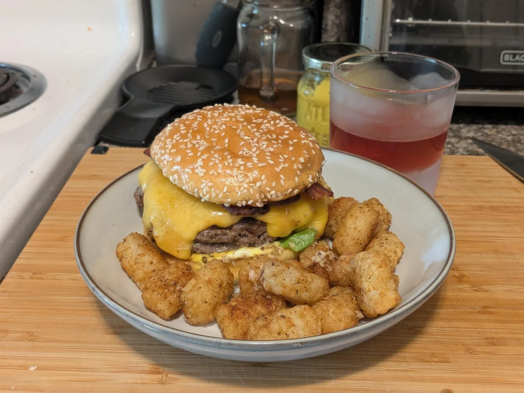 A plate with a double cheeseburger topped with cheddar and bacon, served with tater tots. A glass of iced red drink is beside the plate on a wooden surface in a kitchen setting.