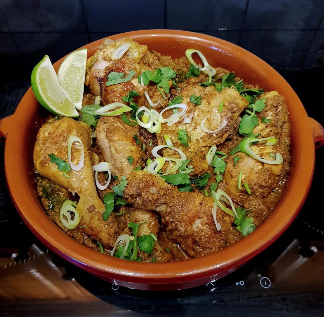 A clay pot filled with seasoned chicken pieces garnished with chopped cilantro, sliced green onions, and lime wedges, placed on a stovetop.