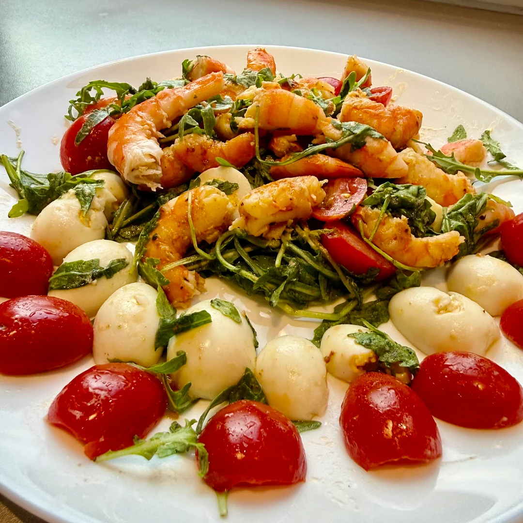 A white plate with shrimp atop arugula and cherry tomatoes, surrounded by mozzarella balls and more cherry tomato halves.