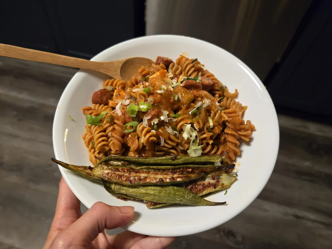 A hand holds a white plate with rotini pasta in sauce, garnished with green onions and cheese. Roasted okra and a wooden spoon are on the side. The background shows a wood floor and dark cabinets.