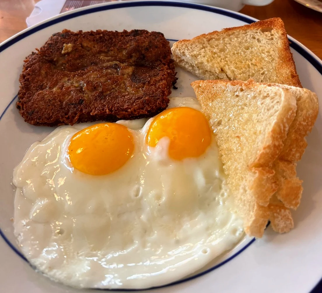 A plate with two sunny-side-up eggs, a crispy brown hash brown patty, and three triangular slices of toasted white bread.