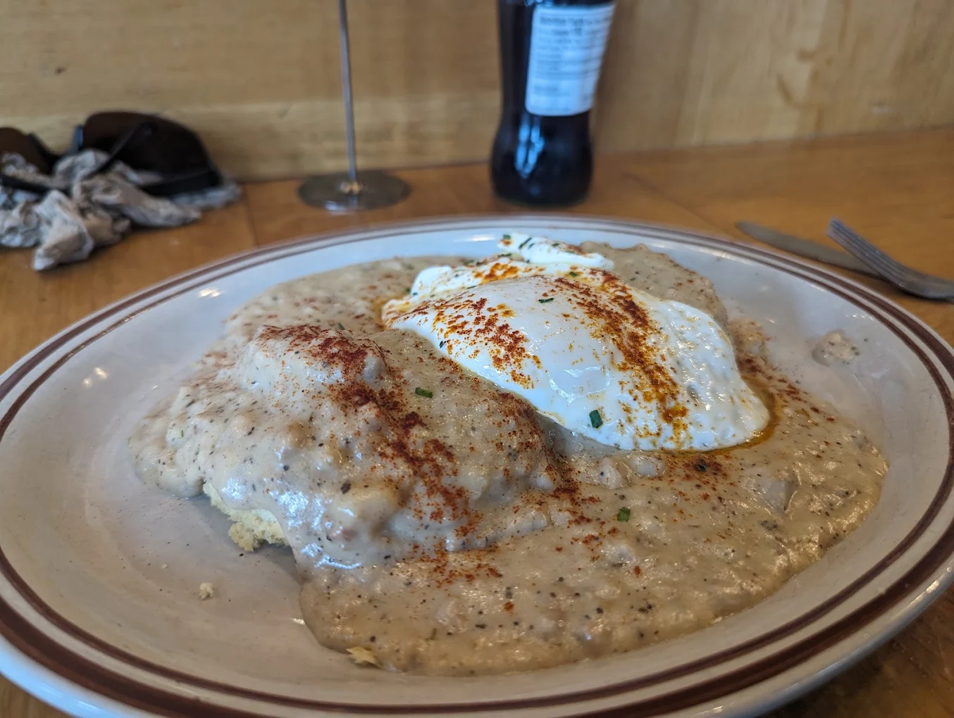 A plate of biscuits and gravy topped with two poached eggs and a sprinkle of paprika, set on a wooden table with a fork, napkin, sunglasses, and a bottled drink in the background.