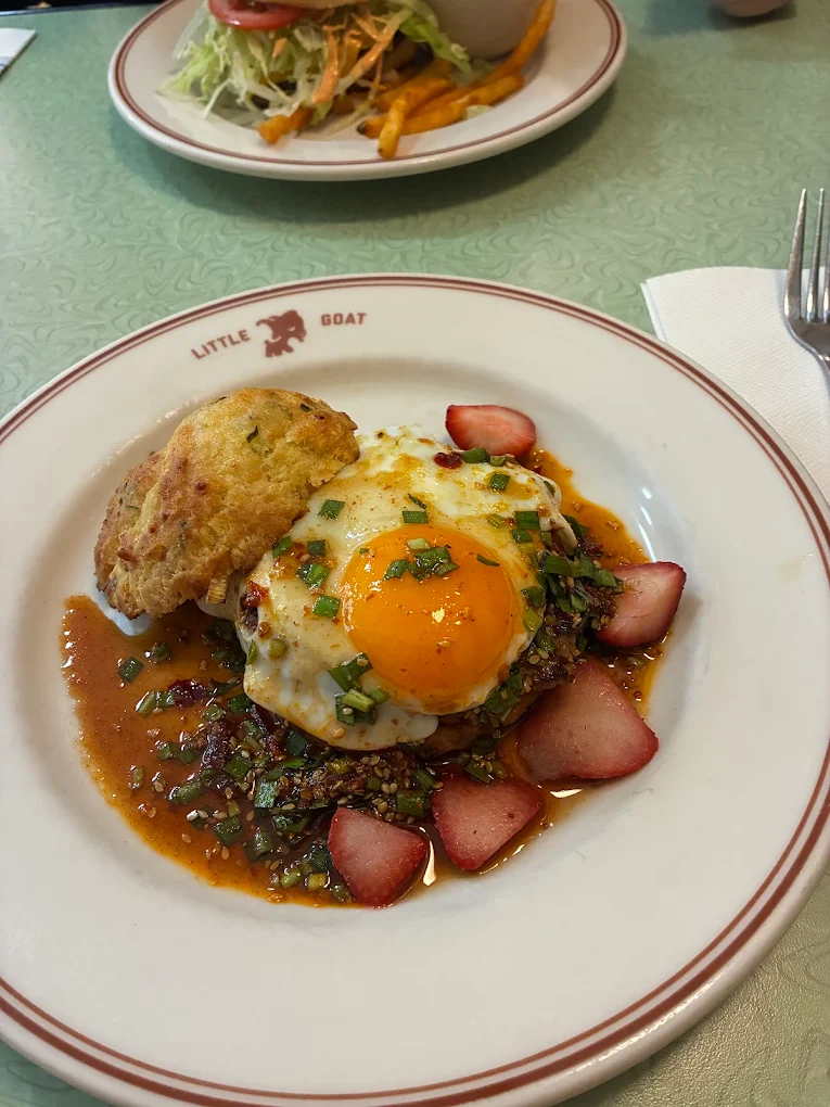 A plate of food featuring a sunny-side-up egg on a patty with sauce, garnished with sliced strawberries and chopped herbs, alongside a biscuit. The plate is labeled "Little Goat." Another dish is visible in the background.