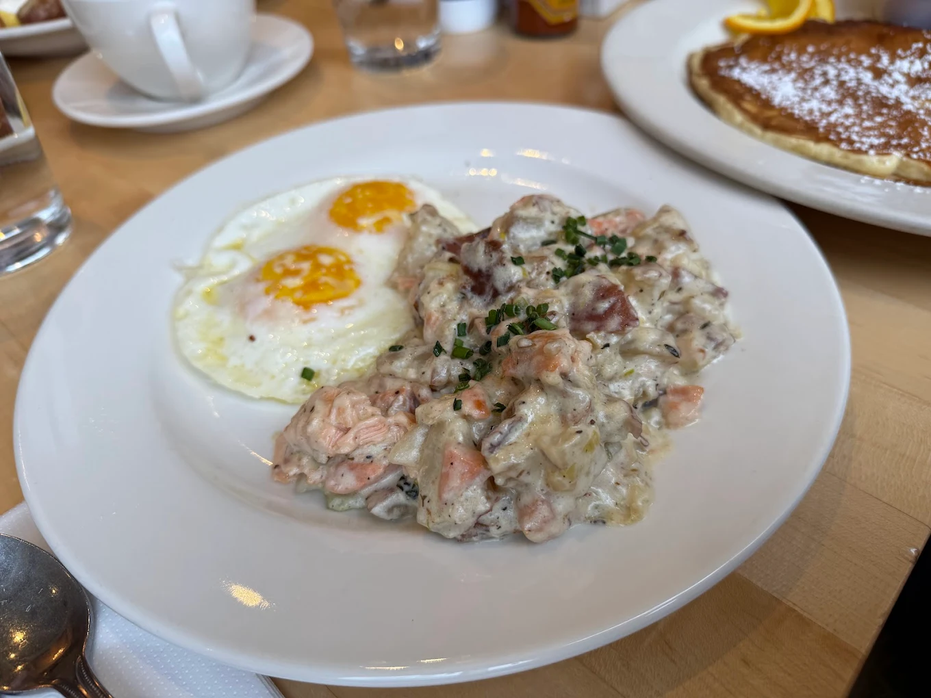 A breakfast plate with two sunny-side-up eggs and a creamy hash with diced meat and chives, with pancakes and orange slices on a separate plate in the background.