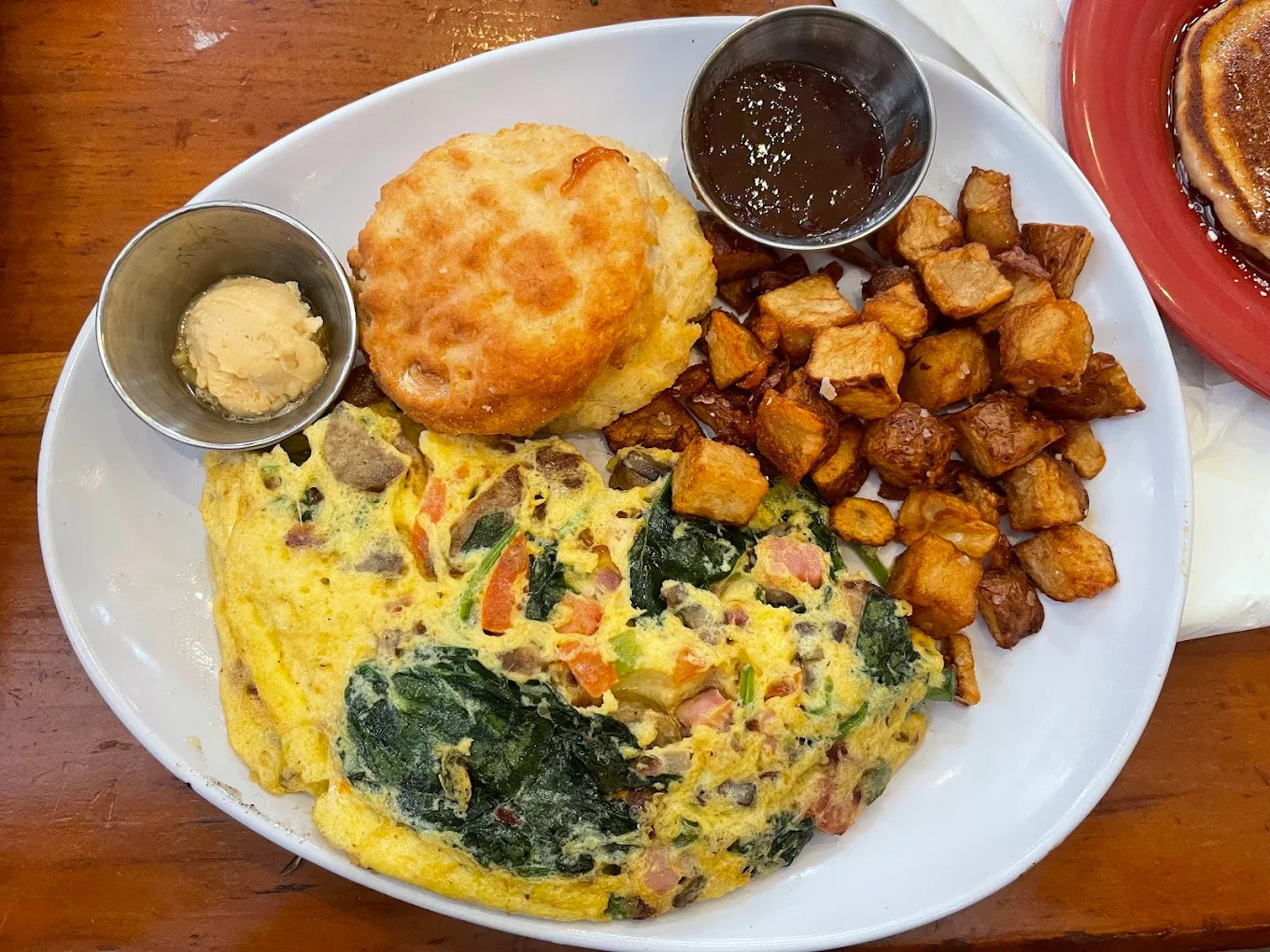 A breakfast plate with a vegetable omelet, a biscuit with a side of butter, crispy diced potatoes, and a small cup of jam, served on a white oval dish on a wooden table.