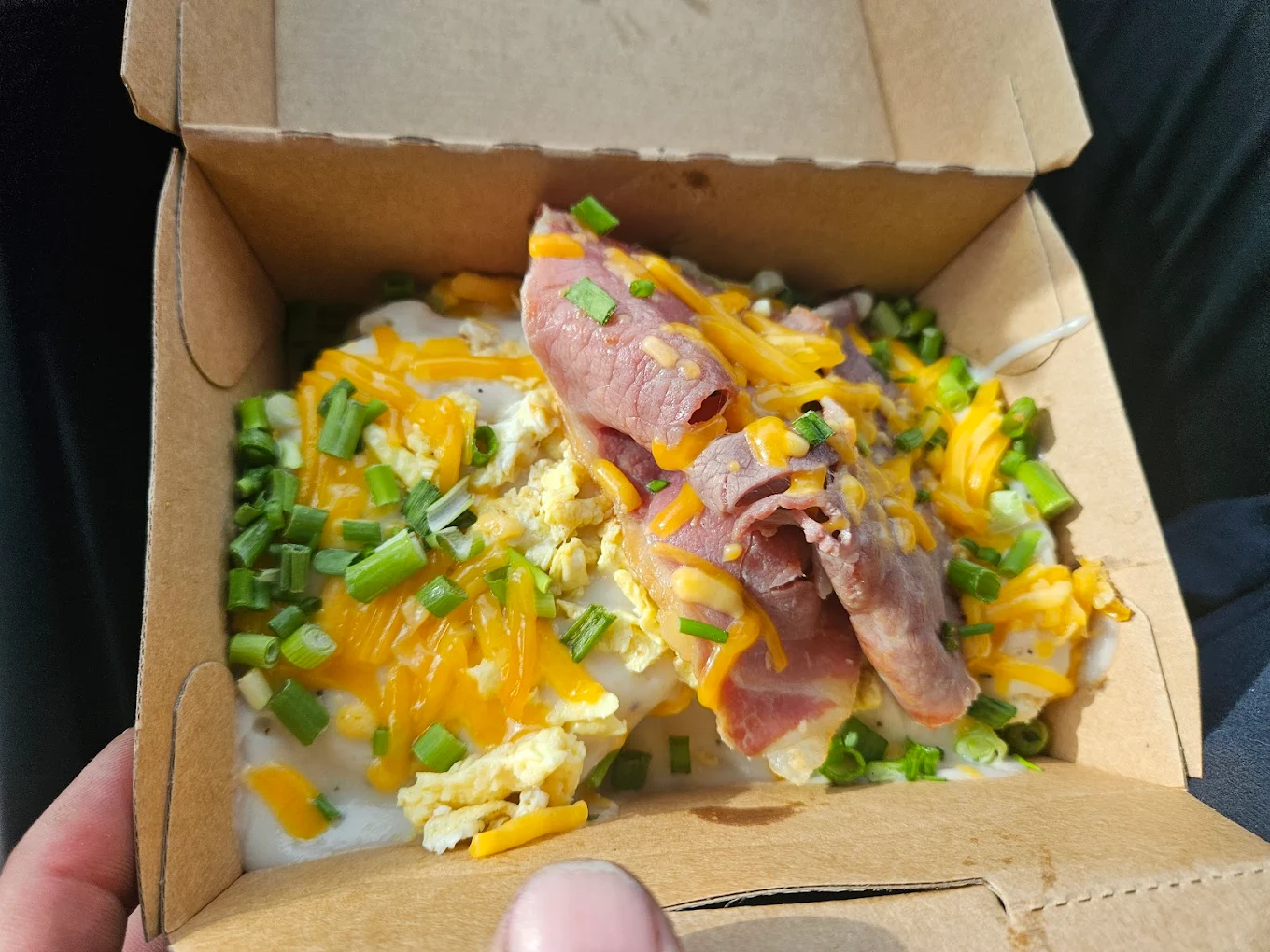 A cardboard takeout box containing scrambled eggs topped with melted cheddar cheese, slices of ham, chopped green onions, and a creamy white sauce. A person's hand is holding the box.