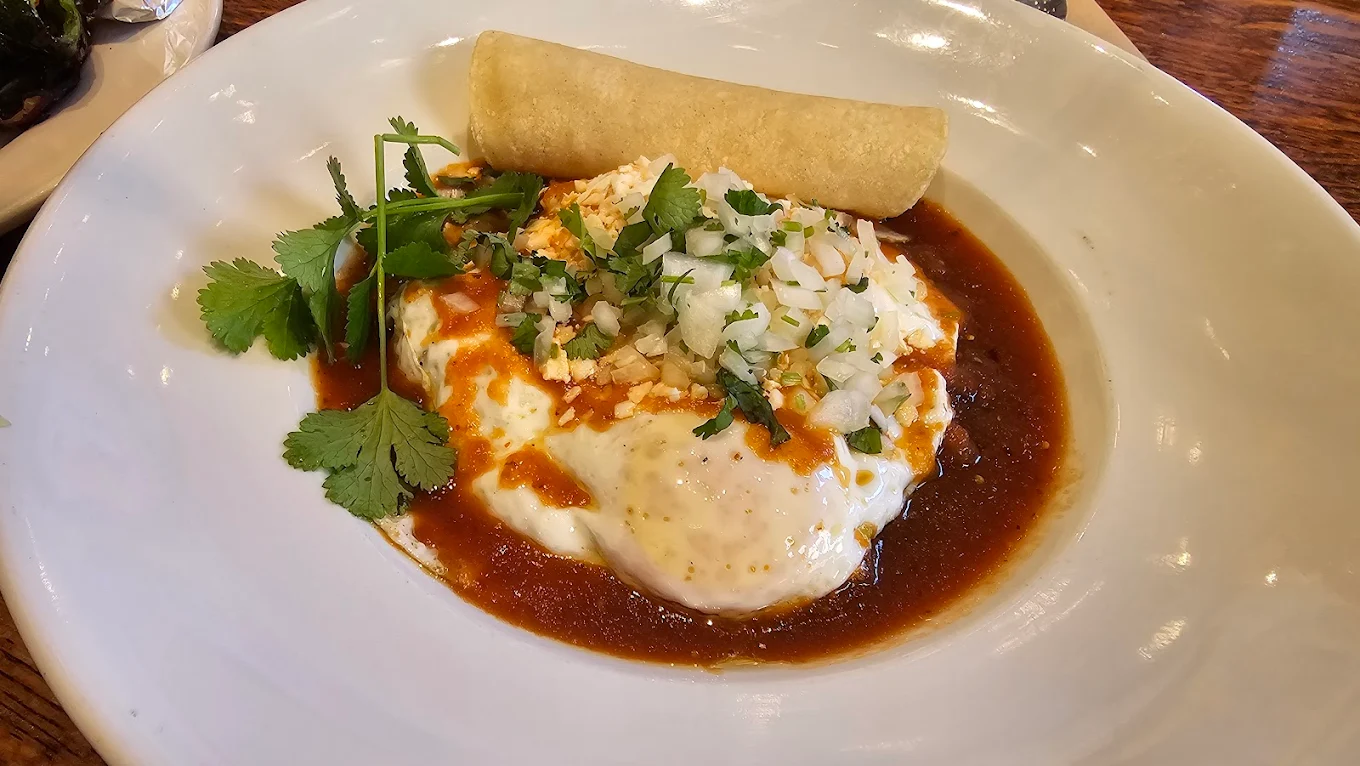 A plate of huevos rancheros topped with chopped onions, cilantro, and cheese, covered in red sauce, with a rolled corn tortilla on the side.