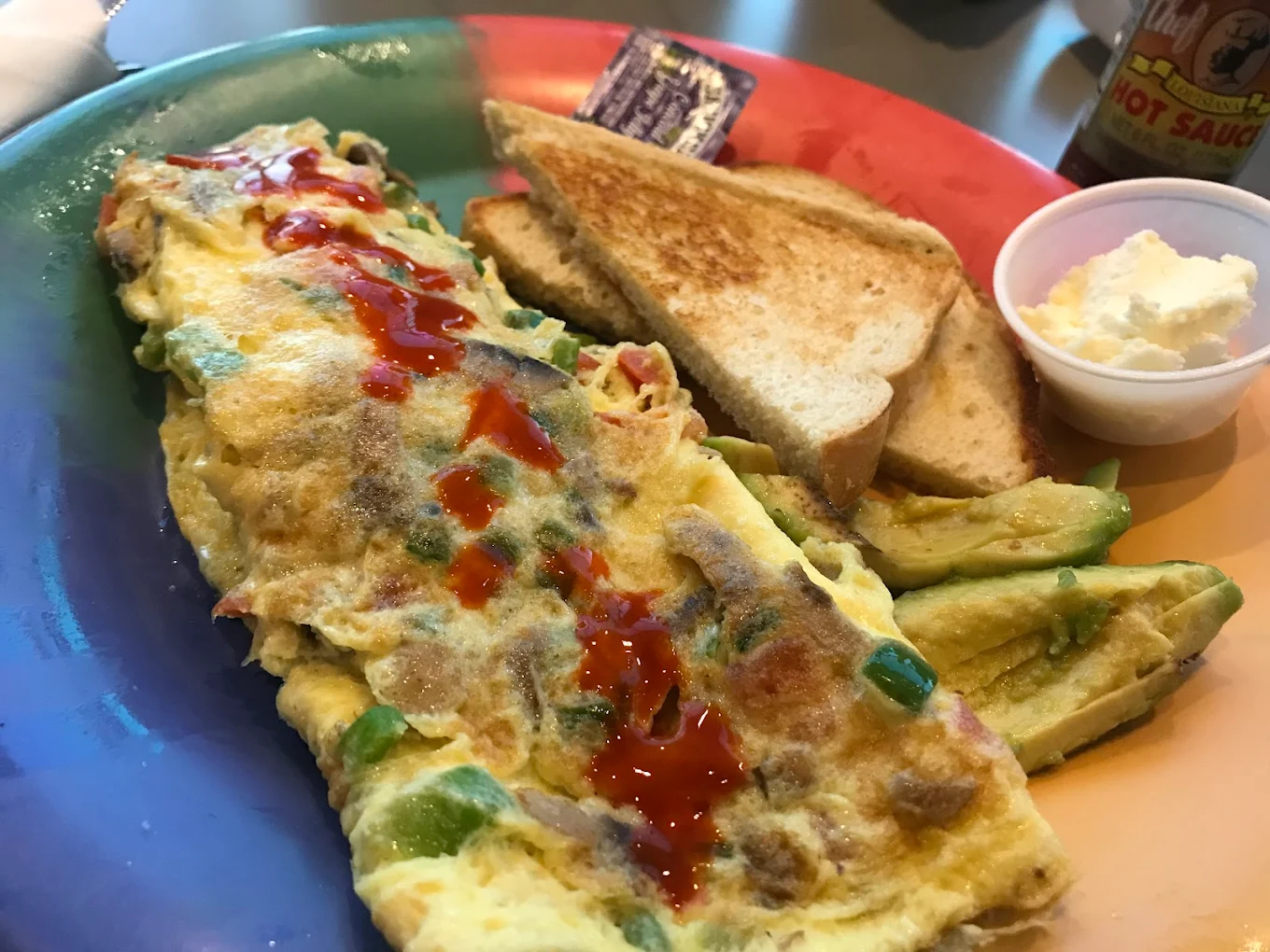 A plate with a vegetable omelette topped with hot sauce, sliced avocado, two pieces of toast, a packet of jelly, and a small cup of whipped butter. Best egg dishes.
