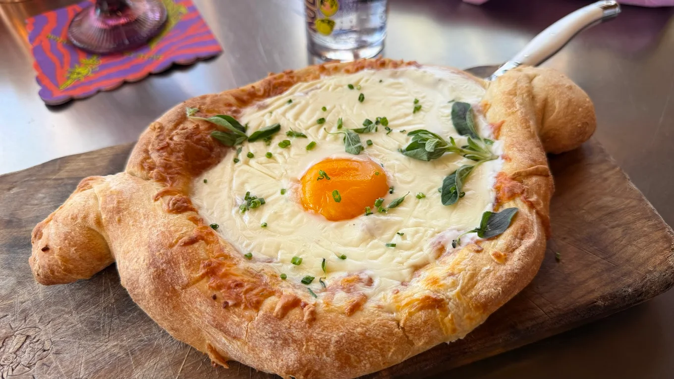 A freshly baked khachapuri, a boat-shaped Georgian bread filled with melted cheese, a sunny egg yolk in the center, and garnished with herbs, sits on a wooden board with a knife beside it.
