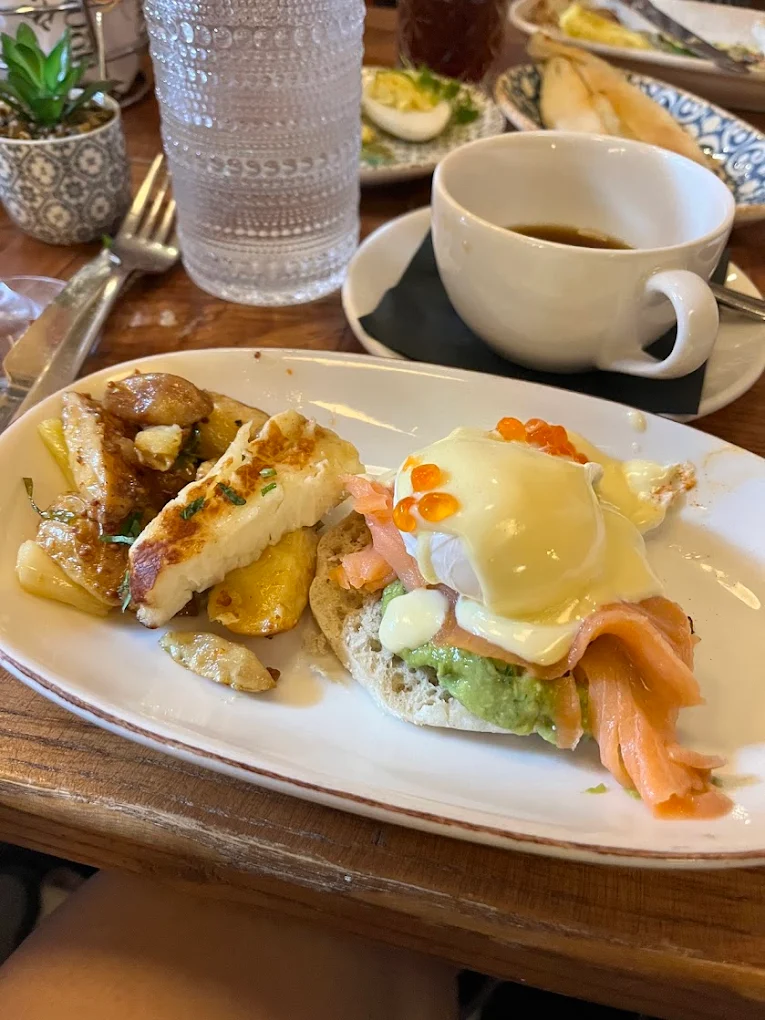 A plate with eggs Benedict topped with hollandaise sauce, smoked salmon, avocado, and salmon roe, served with cheesy potatoes and grilled halloumi; a cup of coffee, cutlery, and a glass of water are beside the plate.