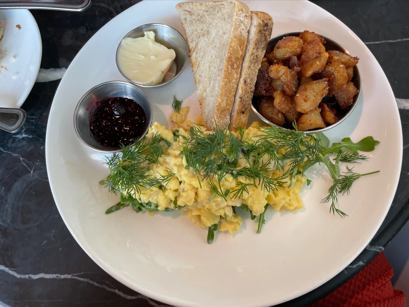A plate with scrambled eggs topped with fresh dill, toast slices, crispy roasted potatoes, a small cup of jam, and a pat of butter, arranged on a white plate on a dark marble table.