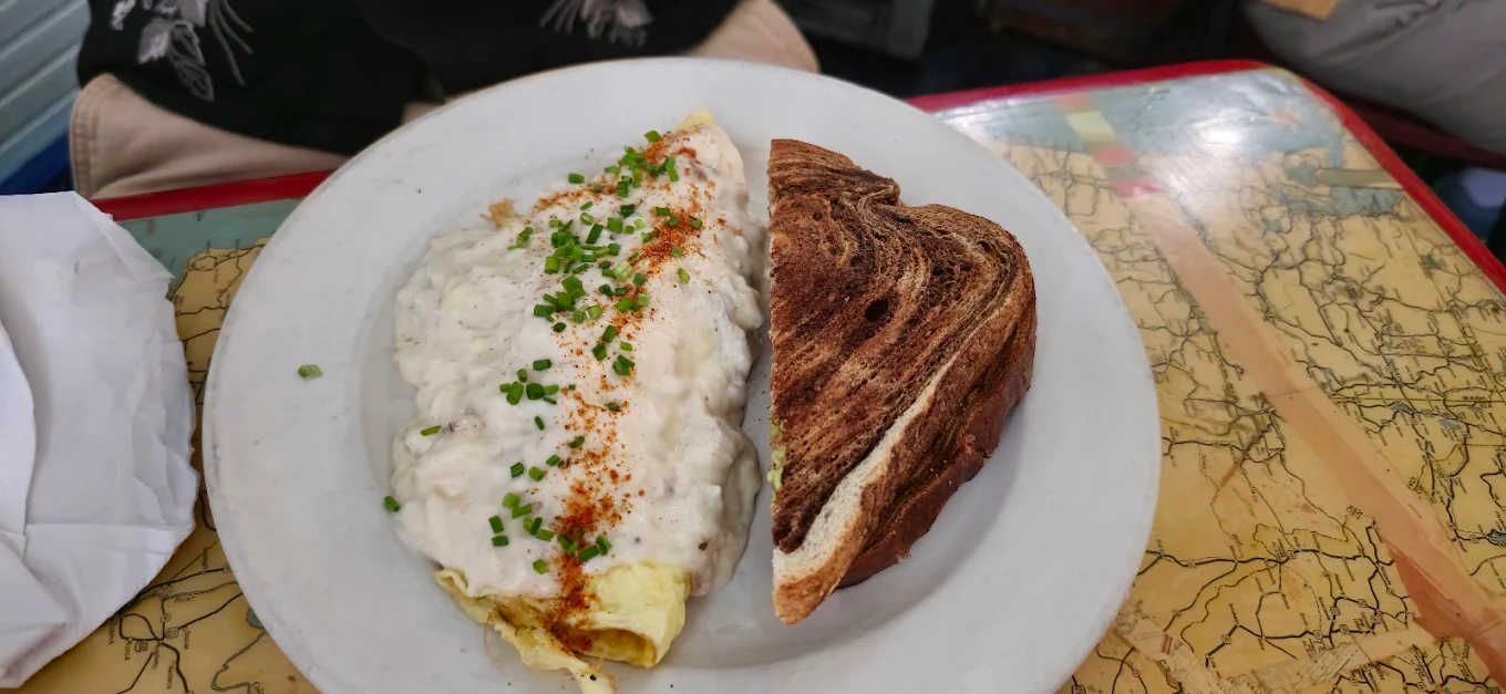 A plate with a folded omelette topped with creamy white sauce, chopped chives, and paprika, served alongside two slices of dark swirled rye bread. The plate is on a colorful, map-patterned table.