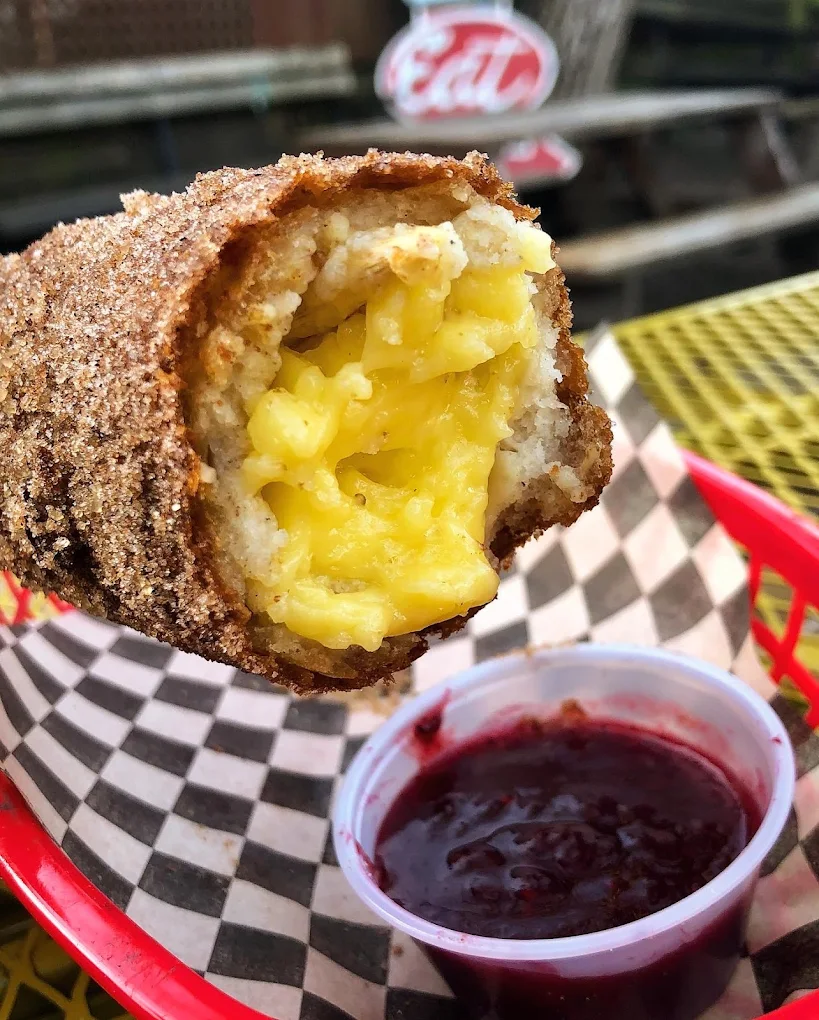A close-up of a deep-fried pastry filled with scrambled eggs, held above a basket lined with checkered paper, next to a small cup of red dipping sauce.