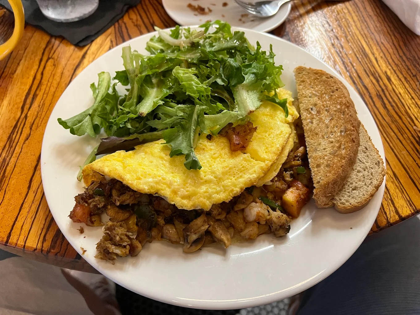 A plate with a folded omelette stuffed with vegetables and potatoes, topped with fresh mixed greens, and two slices of whole grain toast on the side, placed on a wooden table.