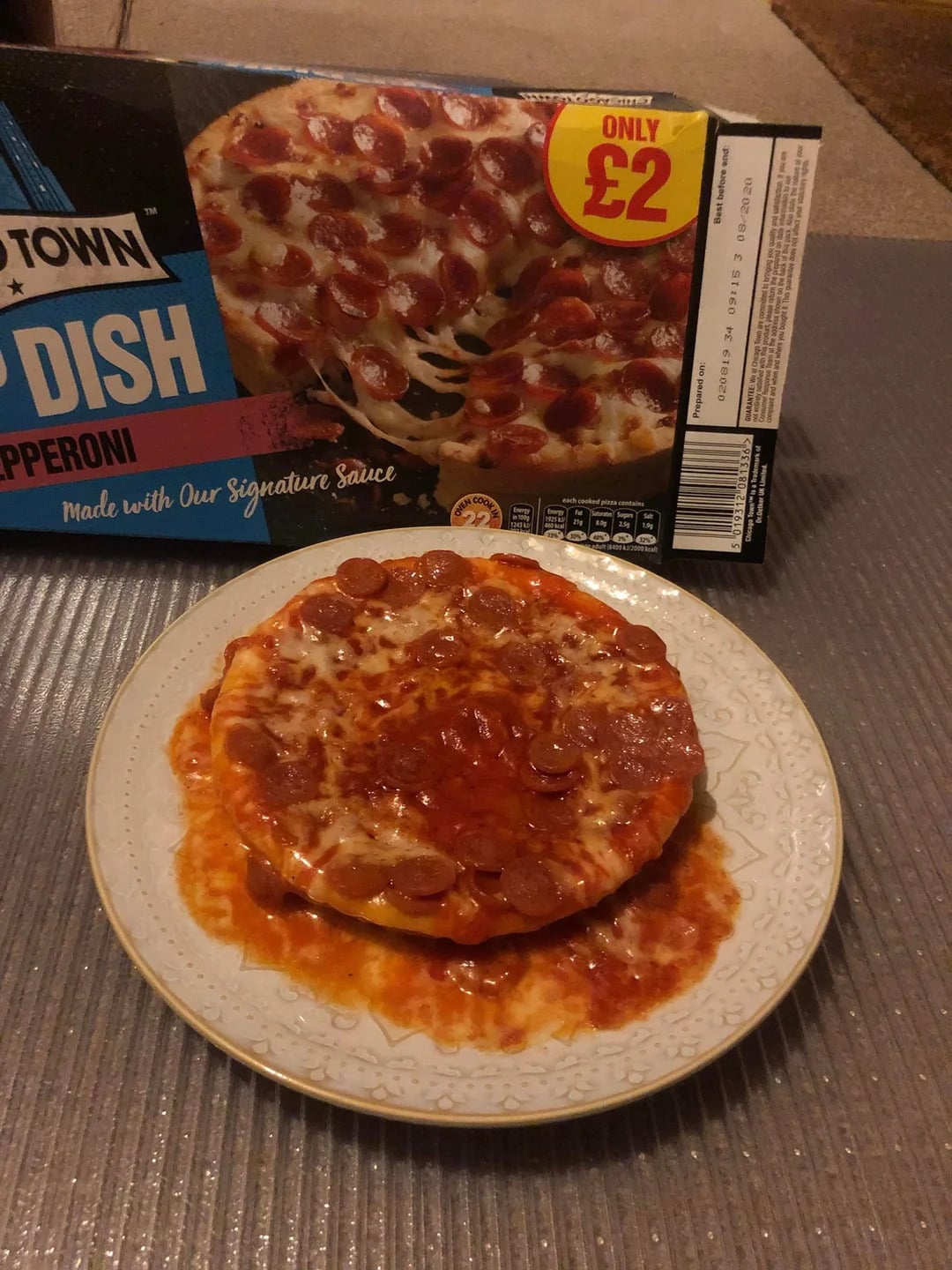 A cooked pepperoni pizza sits on a plate in front of its packaging, which advertises it as "Deep Dish Pepperoni" for £2. The pizza appears smaller and messier than the image on the box.