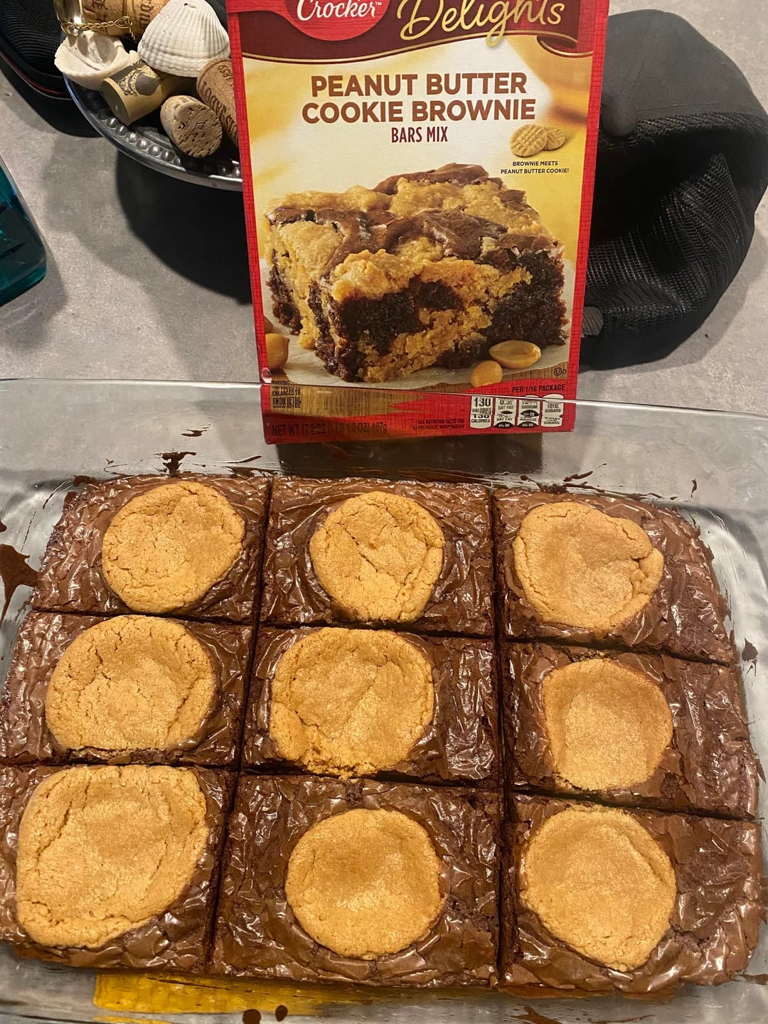 A glass dish of nine baked peanut butter cookie brownies sits in front of a Betty Crocker Peanut Butter Cookie Brownie Bars Mix box on a kitchen counter.