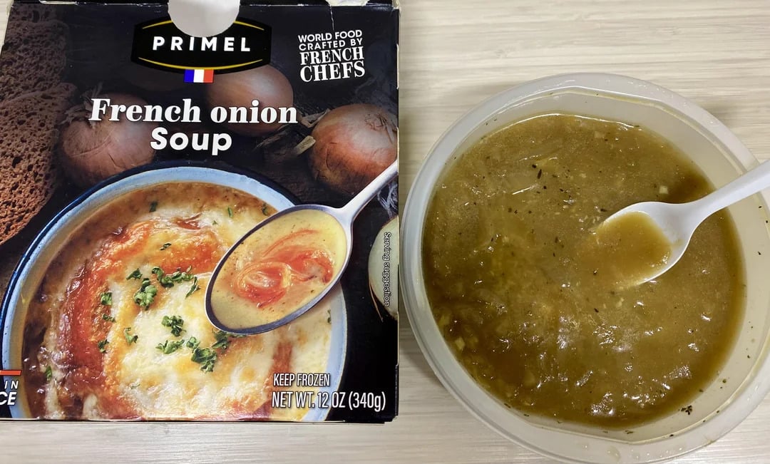 Box of Primel French onion soup next to a bowl of the soup with a plastic spoon. The soup appears brown with herbs, and the packaging shows an image of cheesy, baked French onion soup.