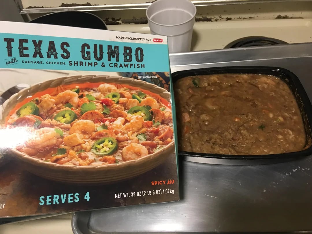 A box of H-E-B Texas Gumbo sits next to the cooked meal in a black tray. The box shows a vibrant gumbo, while the actual dish appears brown and less colorful. Both are on a stovetop.