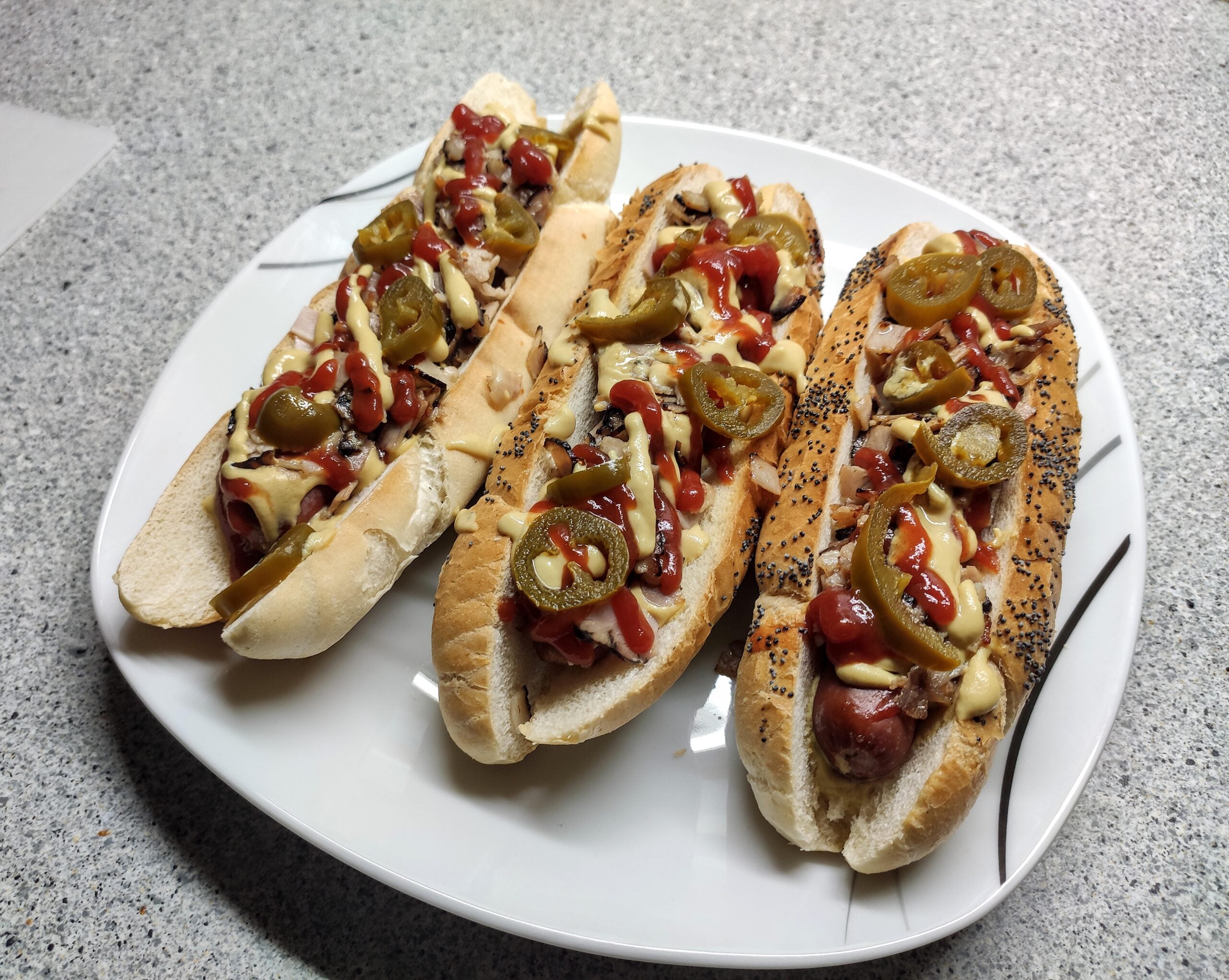 Three hot dogs on a white plate, topped with ketchup, mustard, grilled onions, and sliced jalapeños. The buns include both plain and seeded varieties. The plate is on a speckled countertop.