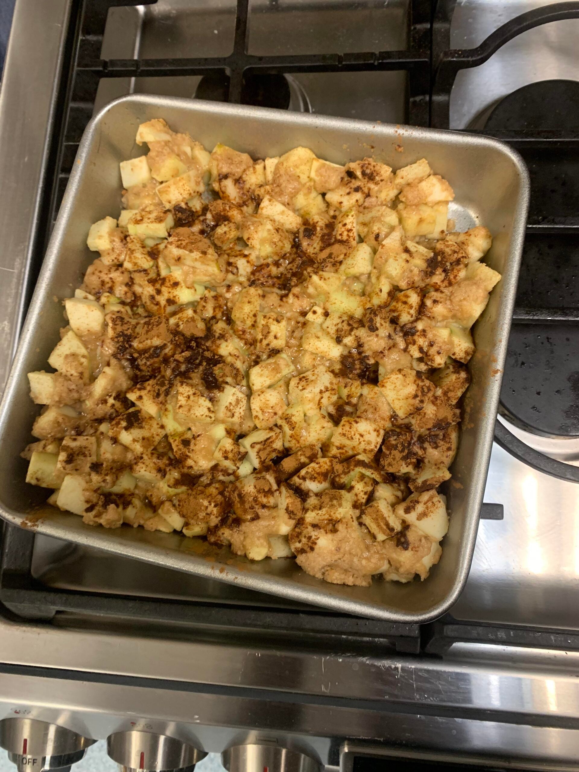 A square metal baking pan filled with chopped apples and a crumb topping sits on a stove, ready to be baked or freshly baked. The topping appears to have cinnamon and brown sugar—a comforting sight that’s worlds away from those hilarious cooking disasters we’ve all experienced in the kitchen.